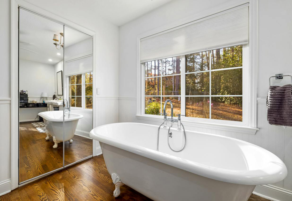 White bathroom with wooden floor, white tub, and window to forest