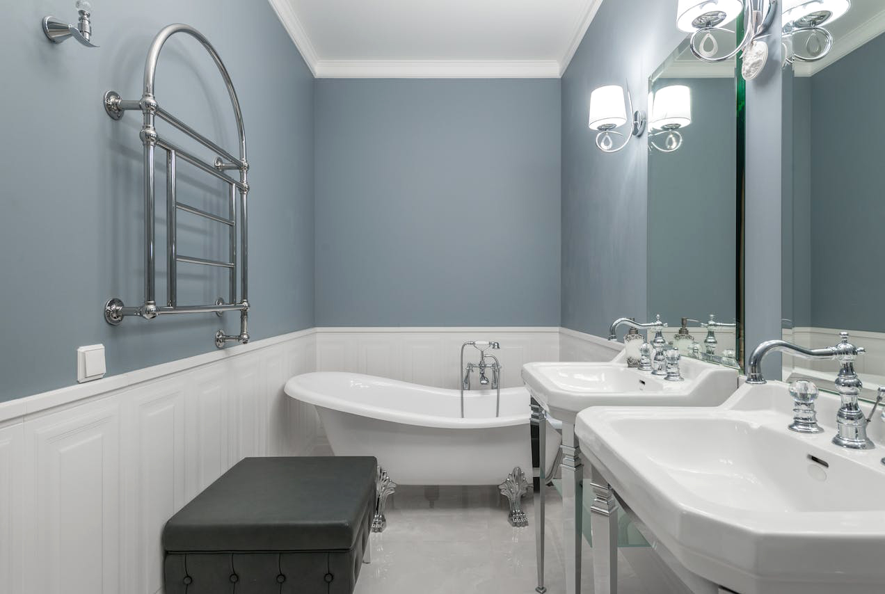 Blueish bathroom with white low wainscoting, white tub, and sinks and a mirrors