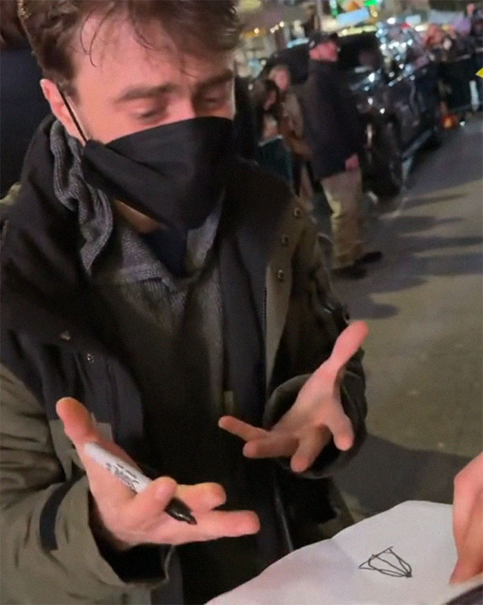 Daniel Radcliffe gesturing with hands, wearing a mask, with a paper showing the Deathly Hallows drawing at an outdoor event. Daniel Radcliffe gesturing with hands, wearing a mask, with a paper showing the Deathly Hallows drawing at an outdoor event.