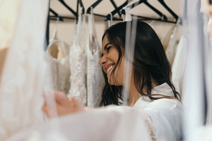 Woman smiling while choosing a wedding dress, surrounded by various lace and embroidered gowns on hangers. Woman smiling while choosing a wedding dress, surrounded by various lace and embroidered gowns on hangers.
