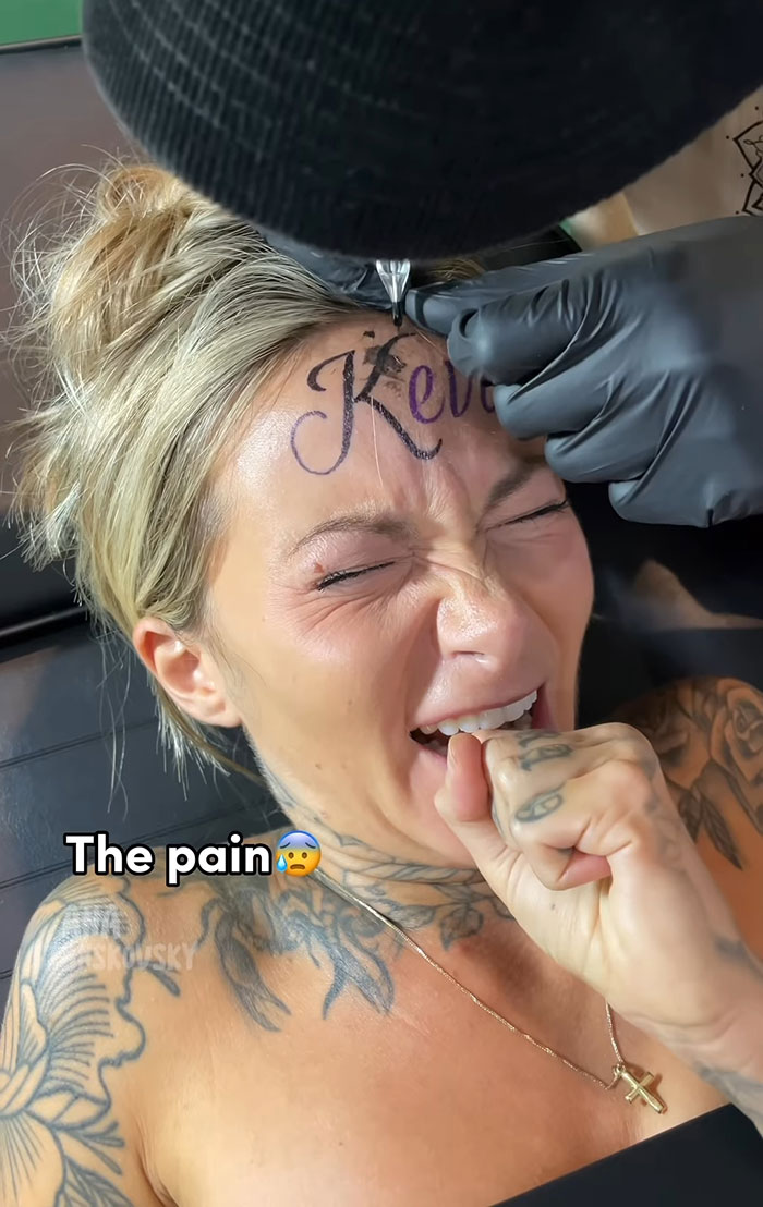 Influencer getting fake tattoo of boyfriend's name on forehead, wincing in pain, tattoo artist in black gloves. Influencer getting fake tattoo of boyfriend's name on forehead, wincing in pain, tattoo artist in black gloves.