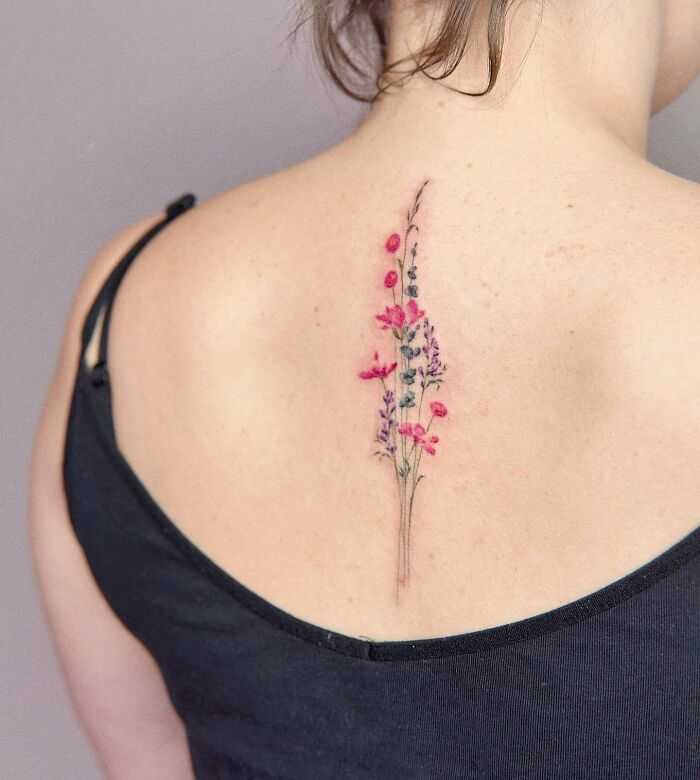 Delicate floral spine tattoo in pink and purple shades on a woman's back.