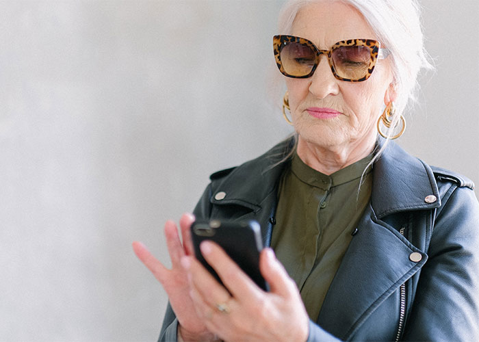 Older woman wearing sunglasses and a leather jacket, focused on her smartphone screen indoors, reacting to paternity test results. Older woman wearing sunglasses and a leather jacket, focused on her smartphone screen indoors, reacting to paternity test results.