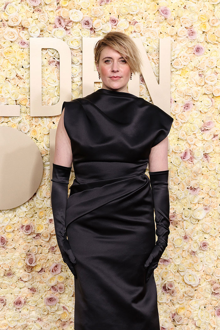 Greta Gerwig in a black dress, standing in front of a floral backdrop at an event. Greta Gerwig in a black dress, standing in front of a floral backdrop at an event.
