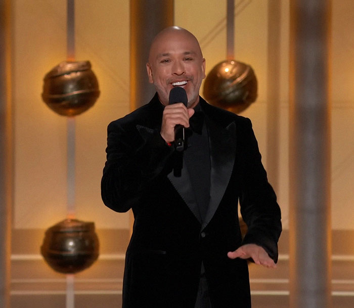 Comedian Jo Koy performs on stage, wearing a black suit, holding a microphone, delivering a joke about Barbie. Comedian Jo Koy performs on stage, wearing a black suit, holding a microphone, delivering a joke about Barbie.