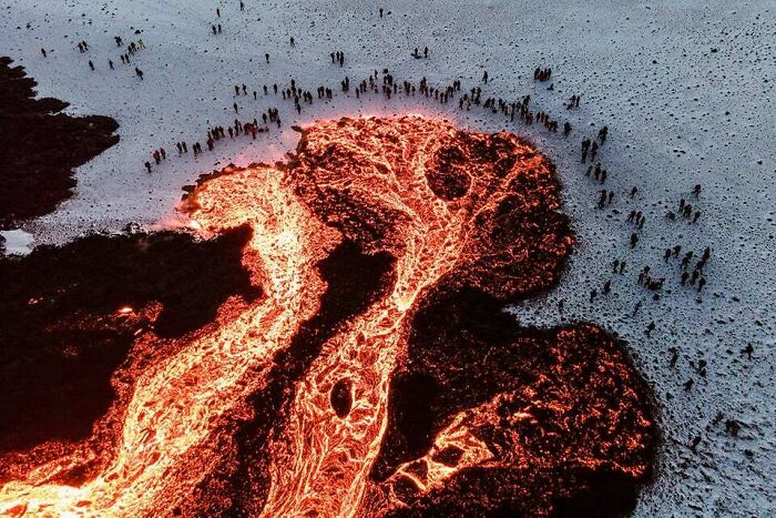 Lava Flowing Across The Snow In Iceland, While A Crowd Of People Warms Themselves Near It While Watching