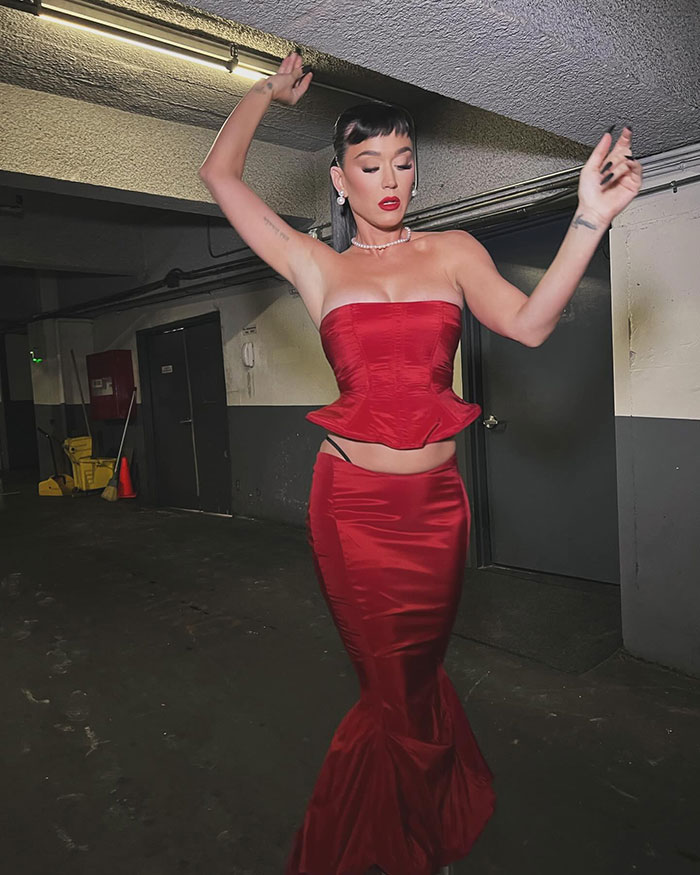 Katy Perry in a red gown showing a black thong and arm tattoo, posing with arms raised. Katy Perry in a red gown showing a black thong and arm tattoo, posing with arms raised.