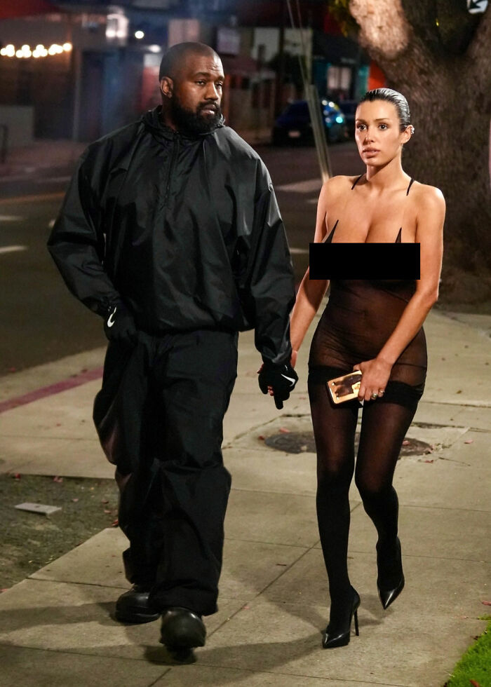 Bianca Censori in a daring outfit, walking on a sidewalk with a man, holding a phone. Bianca Censori in a daring outfit, walking on a sidewalk with a man, holding a phone.