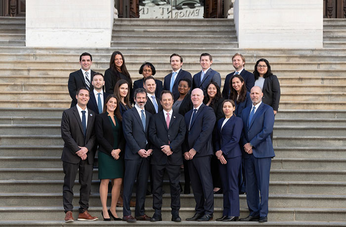 A diverse group of professionals in suits standing on steps, raising awareness about rare uterine cancer affecting 9/11 victims. A diverse group of professionals in suits standing on steps, raising awareness about rare uterine cancer affecting 9/11 victims.