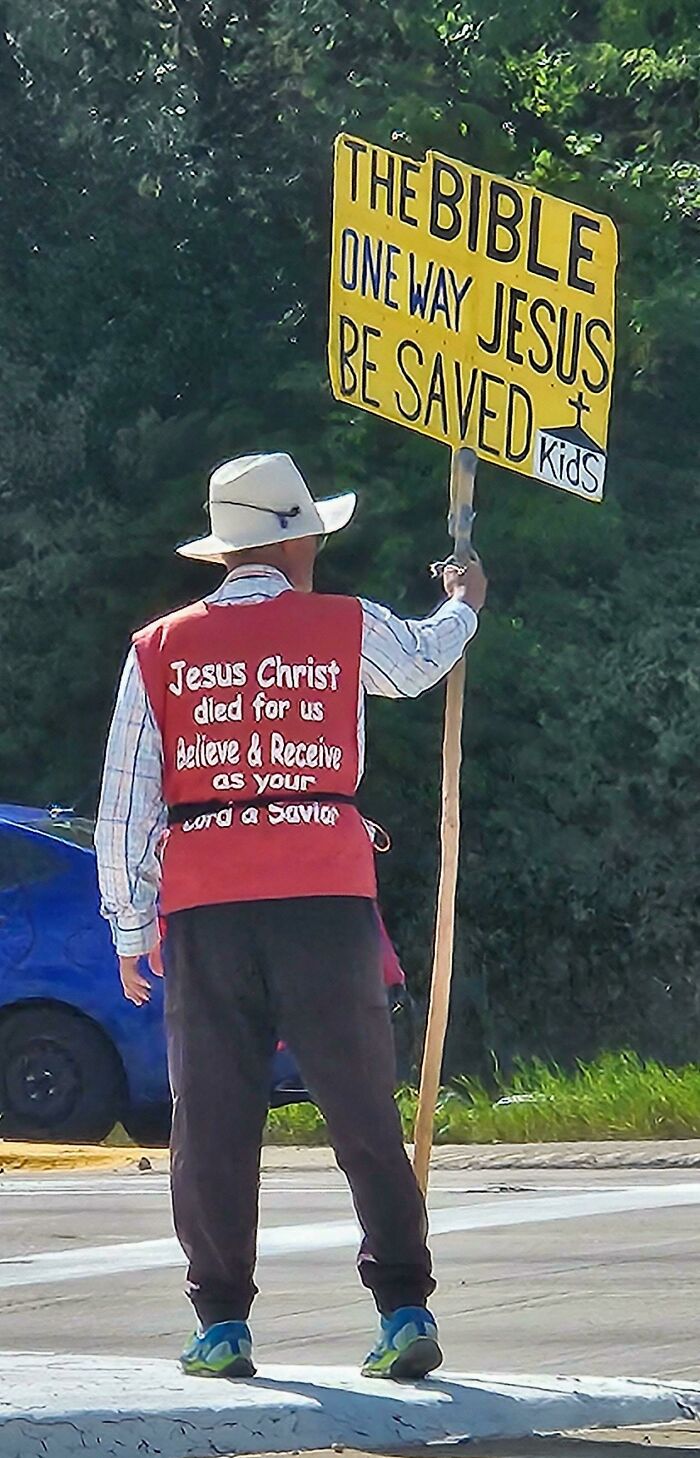 Man holding a religious sign with unintended message, wearing a red vest and hat on a roadside. Sign fails SEO concept.