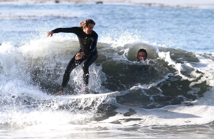 Surfer riding a wave with another person’s head perfectly emerging from the water, showcasing wild coincidences.