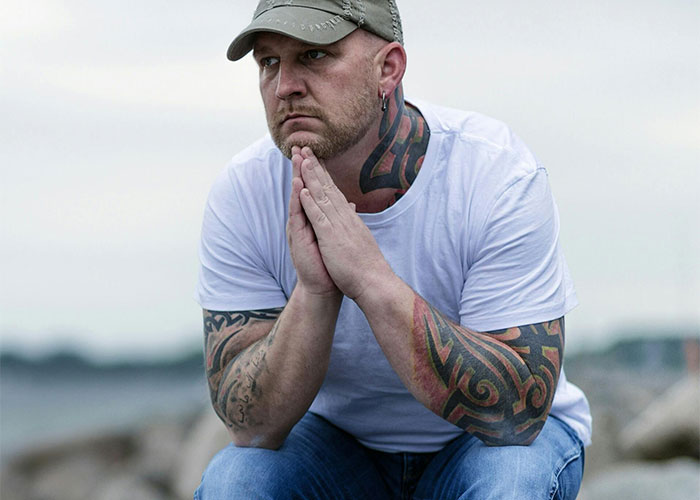 Man with tattoos sits pensively by the sea, wearing a white shirt and cap. Man with tattoos sits pensively by the sea, wearing a white shirt and cap.