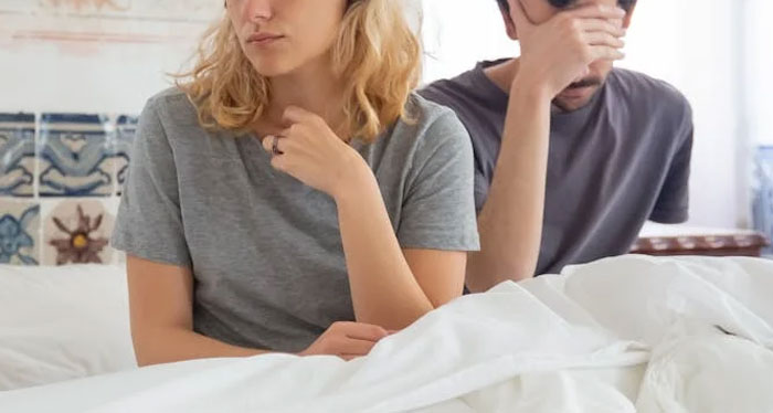 A couple sitting on a bed, woman looking upset and man with hand on his face, upset after a surprise weekend. A couple sitting on a bed, woman looking upset and man with hand on his face, upset after a surprise weekend.
