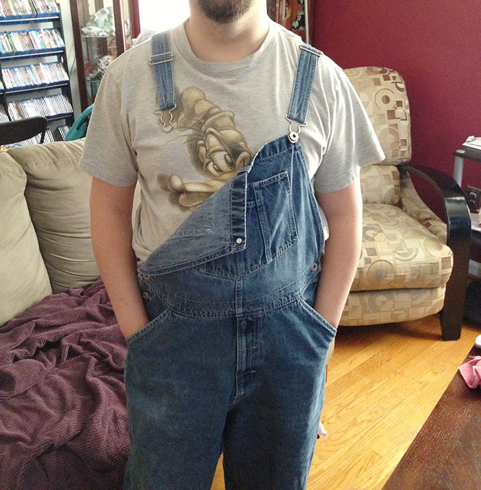 Person wearing denim overalls with one strap undone, sitting in a living room, reflecting millennial trends like whale tail fashion.