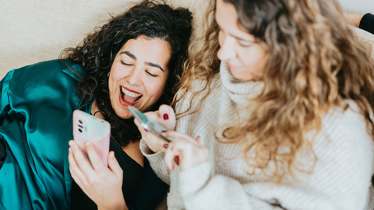 Two women relaxing on a couch, sharing humour and laughing while looking at their smartphones together.