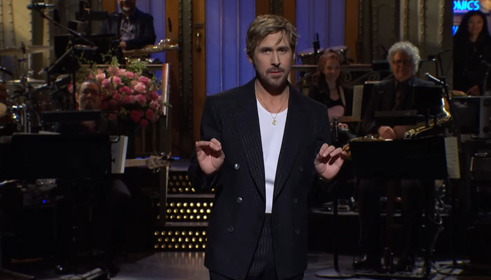 Actor on stage in a dark suit, hands gesturing, with a band and floral arrangement in the background. Actor on stage in a dark suit, hands gesturing, with a band and floral arrangement in the background.