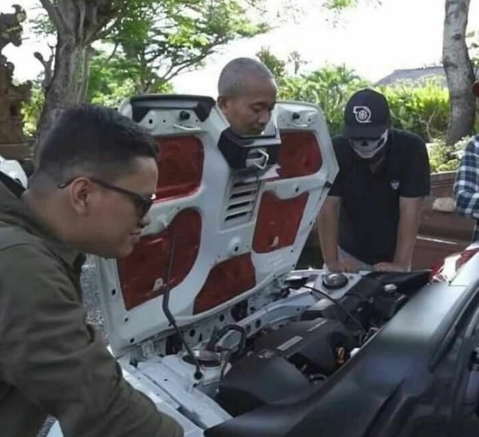 Three men inspecting a car engine with one man humorously wearing a car hood, showcasing a random and cringe moment.
