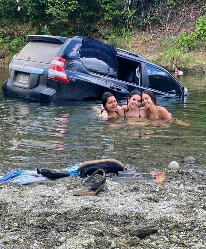 Three women smiling in water near a partially submerged black SUV in a random and cringe photo with unique humor.