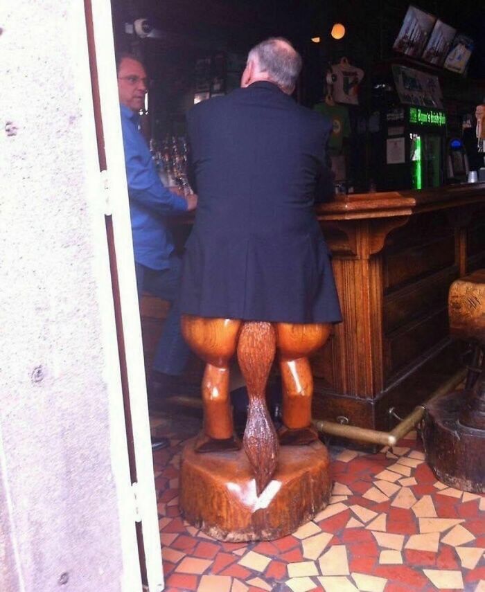 Man sitting on a random and cringe wooden stool shaped like animal legs inside a dimly lit bar.