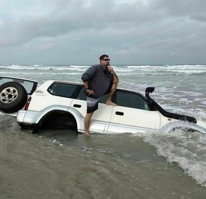 Man posing on half-sunken white SUV in ocean waves, a random and cringe photo for those with unique sense of humor.