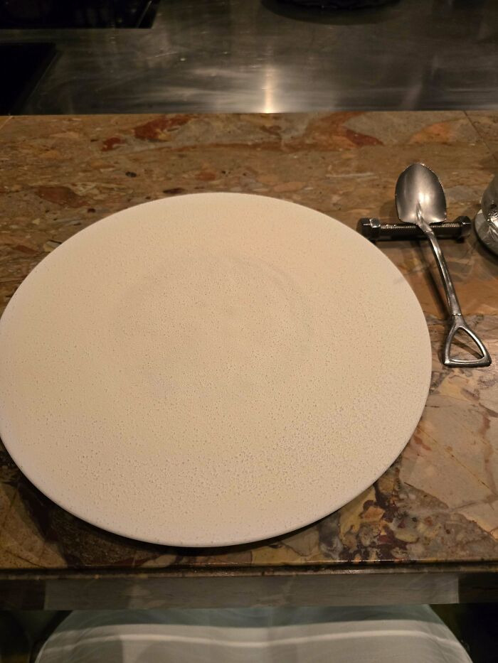 Empty textured plate with a small shovel-shaped spoon on a marble tabletop showing ridiculous ways food served in restaurants.