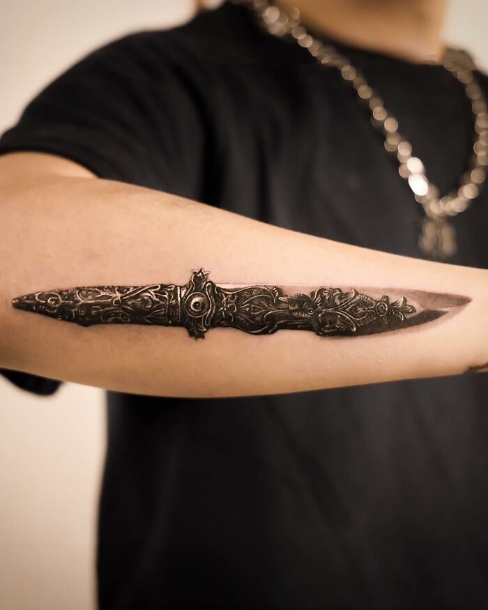 Hyperrealistic tattoo of a detailed dagger on a person's forearm, showcasing intricate design and shading. Hyperrealistic tattoo of a detailed dagger on a person's forearm, showcasing intricate design and shading.