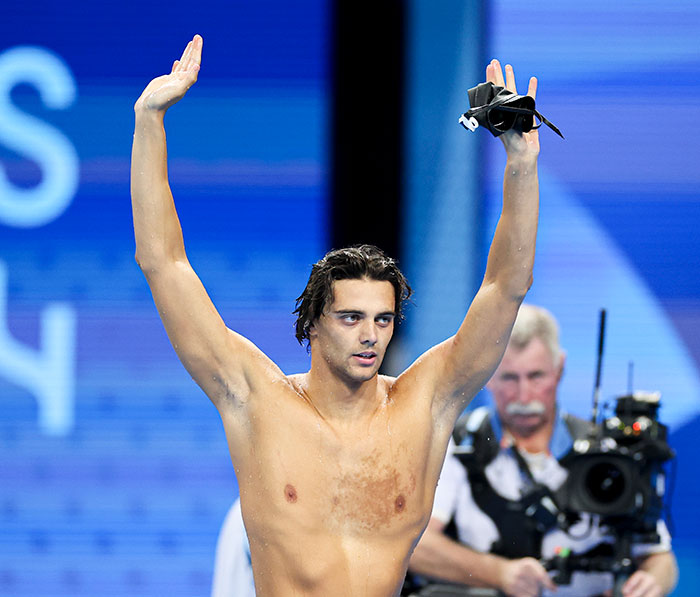 Swimmer celebrating a victory with arms raised, wearing swimming goggles in hand, surrounded by media. Swimmer celebrating a victory with arms raised, wearing swimming goggles in hand, surrounded by media.