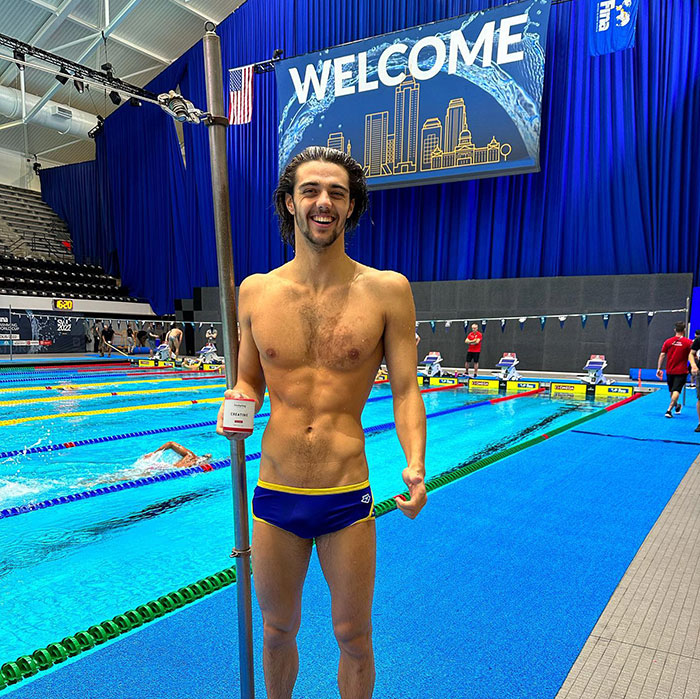 Gold medalist in swim briefs at a pool event, smiling and holding onto a pole, with a blue backdrop and welcome sign. Gold medalist in swim briefs at a pool event, smiling and holding onto a pole, with a blue backdrop and welcome sign.