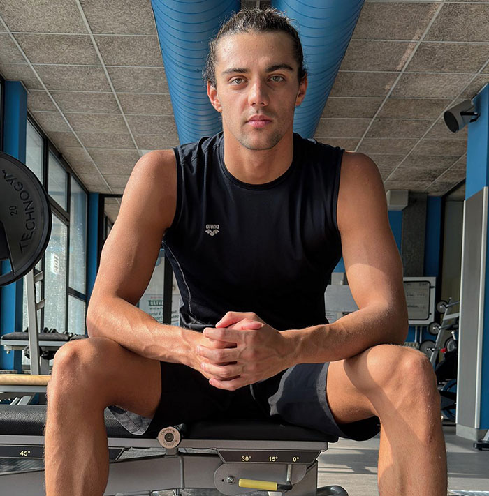 Gold medalist Thomas Ceccon seated in a gym wearing a black tank top, looking at the camera. Gold medalist Thomas Ceccon seated in a gym wearing a black tank top, looking at the camera.