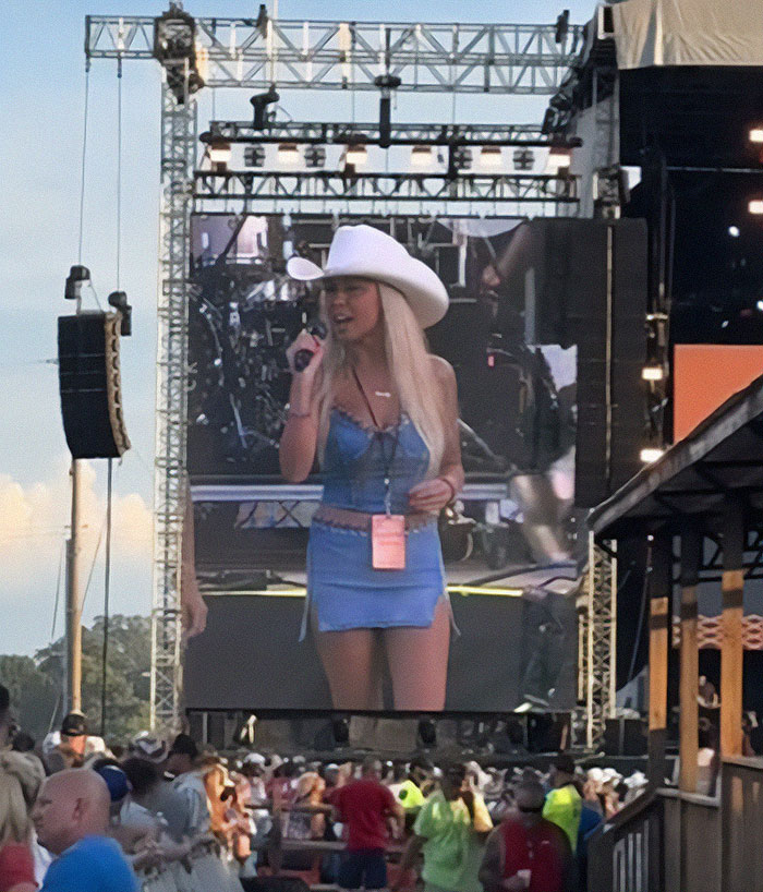 Hailey Welch performing live on stage, wearing a blue dress and white cowboy hat, while concert crowd watches silently. Hailey Welch performing live on stage, wearing a blue dress and white cowboy hat, while concert crowd watches silently.