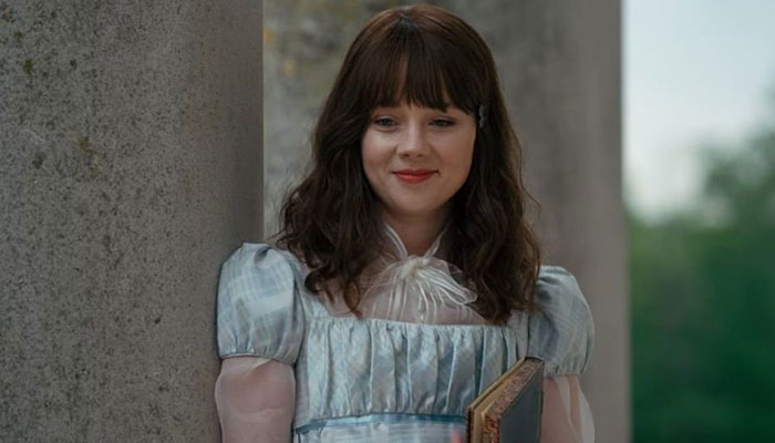 Makeup artist covers up tattoo on actress Claudia Jessie in period dress, standing against a stone pillar, looking content. Makeup artist covers up tattoo on actress Claudia Jessie in period dress, standing against a stone pillar, looking content.