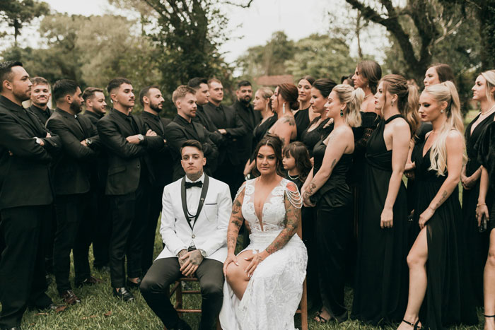 Bride and groom sitting, surrounded by bridesmaids and groomsmen in black attire, outdoors. Bride and groom sitting, surrounded by bridesmaids and groomsmen in black attire, outdoors.