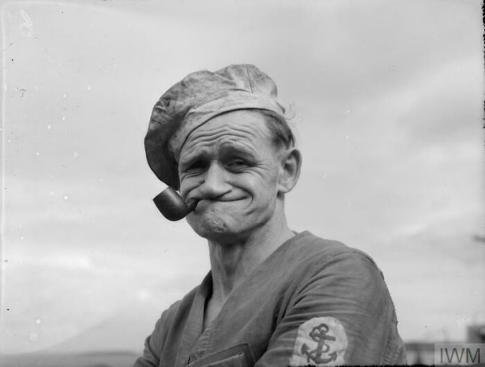 Black and white photo of an older man with creaking joints, wearing a sailor hat and smoking a pipe outdoors.