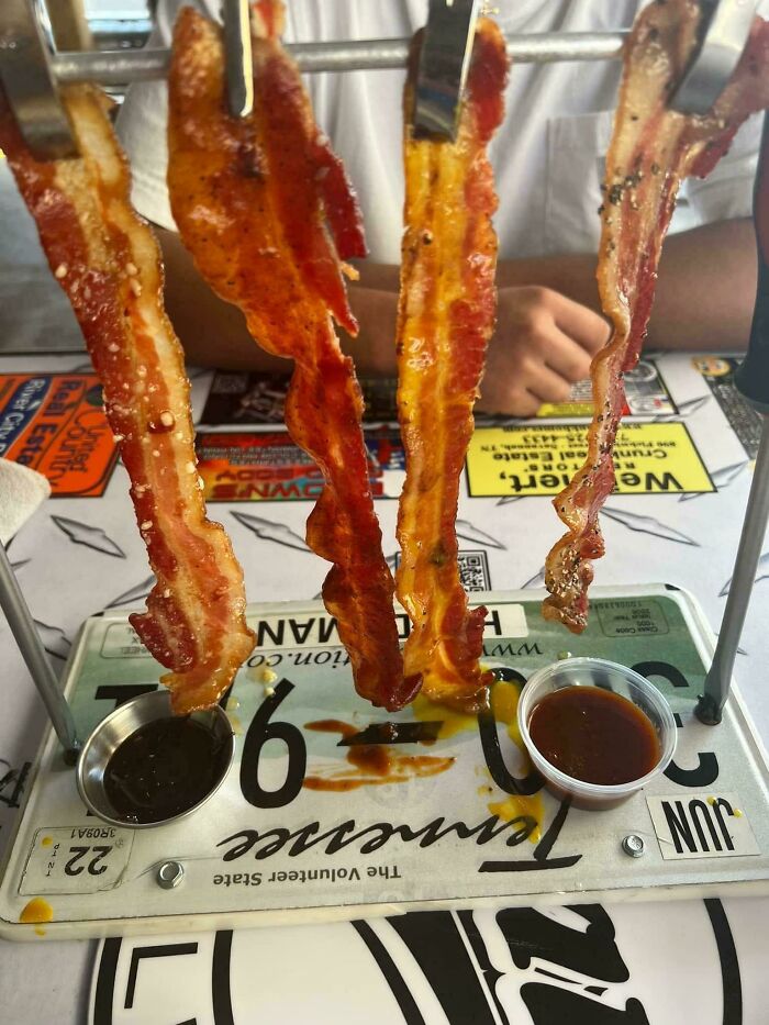 Bacon strips hanging from a metal rack over a license plate with two dipping sauces in a restaurant setting.