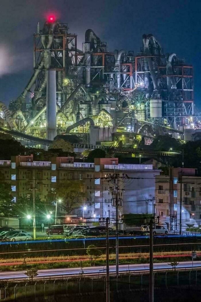 Large industrial factory complex lit up at night towering over residential buildings, evoking megalophobia fears.