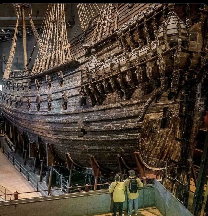 Massive old wooden ship exhibited indoors towering over visitors, evoking megalophobia with its immense size and detailed structure.