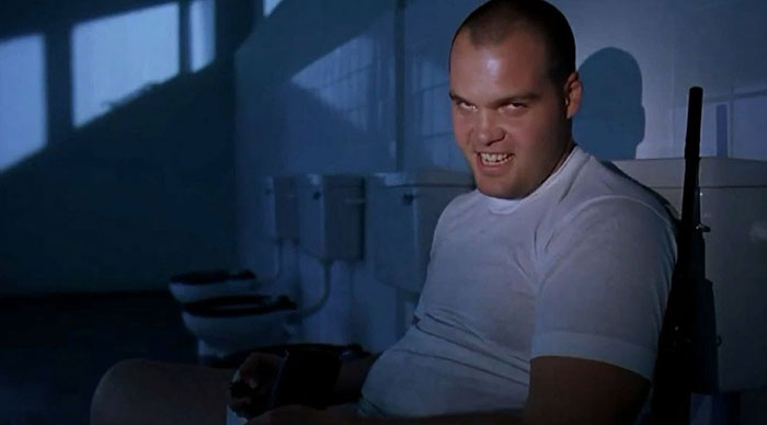 A dimly lit scene featuring a man in a white t-shirt sitting in a bathroom, with a menacing grin on his face. His intense expression, combined with the dark shadows cast in the room, creates an unsettling atmosphere. The bathroom stalls and toilets in the background add to the eerie feeling. A dimly lit scene featuring a man in a white t-shirt sitting in a bathroom, with a menacing grin on his face. His intense expression, combined with the dark shadows cast in the room, creates an unsettling atmosphere. The bathroom stalls and toilets in the background add to the eerie feeling.