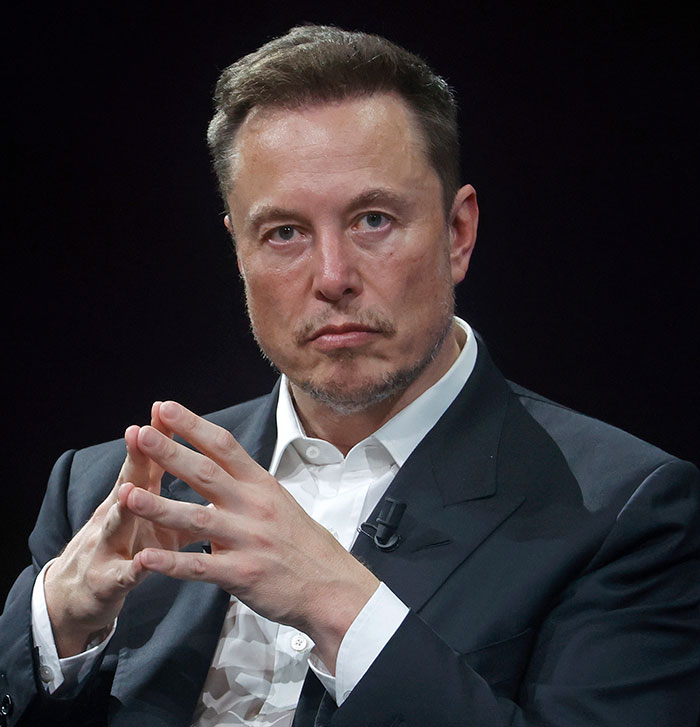 Elon Musk in a dark suit, concentrating with hands clasped during a public event. Elon Musk in a dark suit, concentrating with hands clasped during a public event.