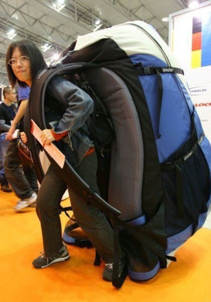 A person wearing an absurdly oversized backpack, which is so large that it dwarfs them, making it appear comically impractical for normal use. The person is leaning forward slightly as if struggling under the weight of the giant pack. A person wearing an absurdly oversized backpack, which is so large that it dwarfs them, making it appear comically impractical for normal use. The person is leaning forward slightly as if struggling under the weight of the giant pack.