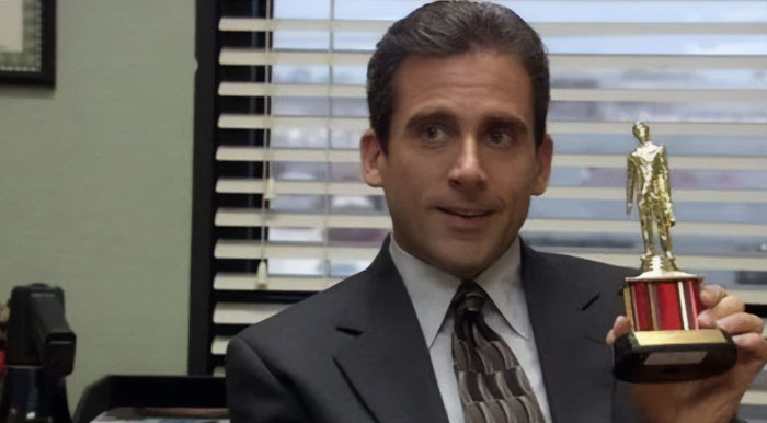 The image shows a character in a suit holding a small trophy with a figure on top, while seated in an office setting with blinds in the background. The character is smiling slightly, as if proud of the trophy. The image shows a character in a suit holding a small trophy with a figure on top, while seated in an office setting with blinds in the background. The character is smiling slightly, as if proud of the trophy.