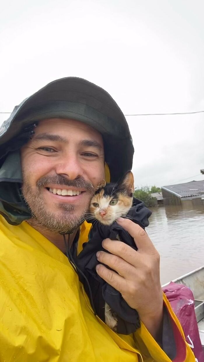 Man in a raincoat smiling, holding a kitten, embodying the hero without a cape spirit amidst floodwaters. Man in a raincoat smiling, holding a kitten, embodying the hero without a cape spirit amidst floodwaters.
