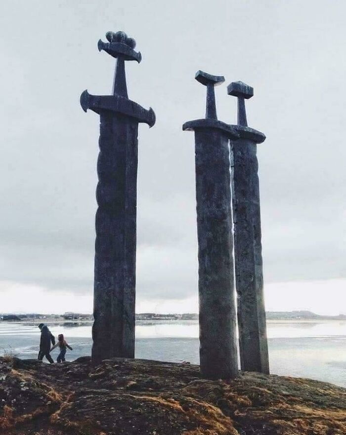 Three giant sword sculptures towering over people near a rocky lakeside, evoking megalophobia fear.