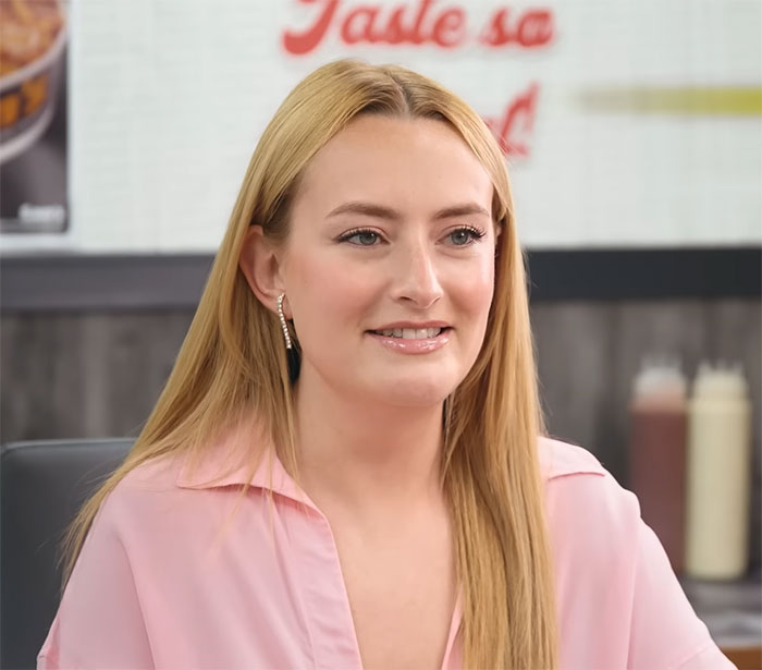 Amelia Dimoldenberg in a pink blouse smiling, with sauce bottles in the background; relates to "Masterclass In Seduction" video theme. Amelia Dimoldenberg in a pink blouse smiling, with sauce bottles in the background; relates to "Masterclass In Seduction" video theme.