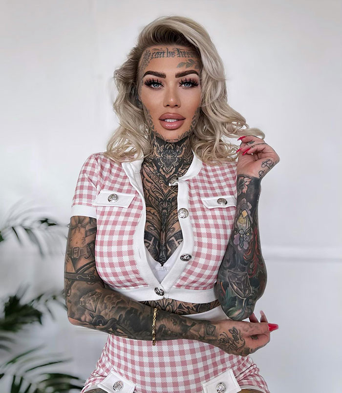 A woman with face tattoos wearing a checkered outfit stands confidently in front of a neutral background. A woman with face tattoos wearing a checkered outfit stands confidently in front of a neutral background.