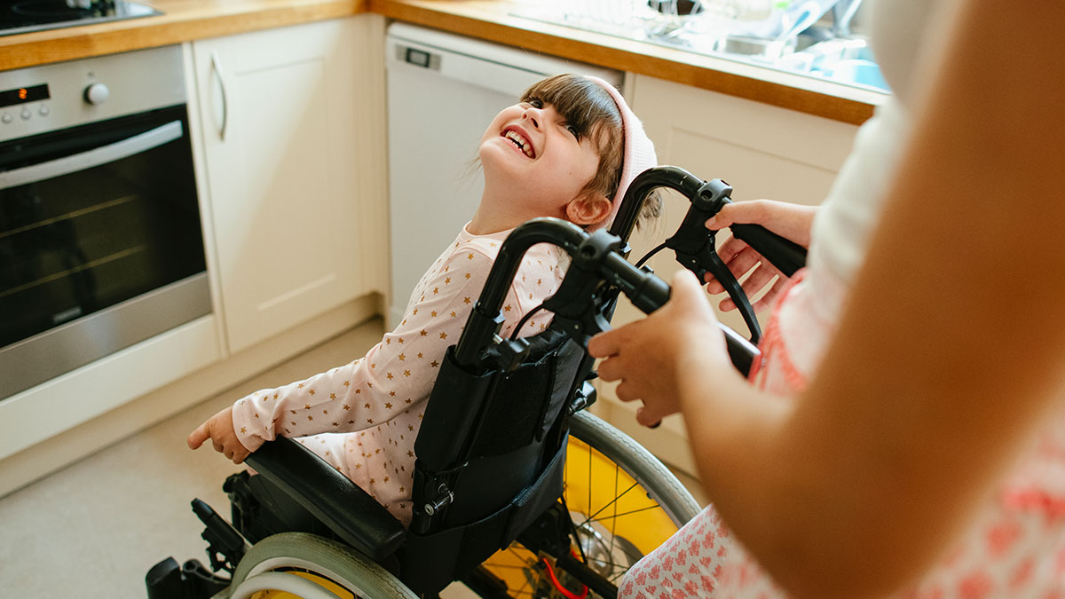 Young woman in wheelchair smiling happily while being pushed through a kitchen during a housewarming party.