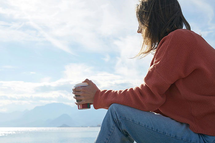 Woman in a red sweater holding a cup, sitting outdoors by water, reflecting after a housewarming party sister fight incident. Woman in a red sweater holding a cup, sitting outdoors by water, reflecting after a housewarming party sister fight incident.