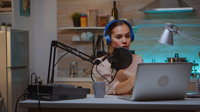 Woman recording a podcast at home, wearing blue headphones, speaking into a microphone, with a laptop open in front. Woman recording a podcast at home, wearing blue headphones, speaking into a microphone, with a laptop open in front.