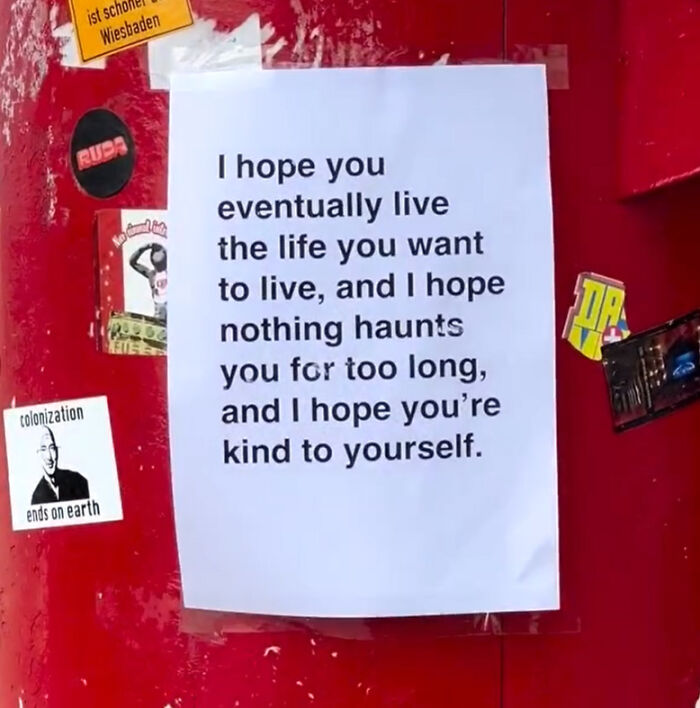White paper with a wholesome message about living the life you want, taped on a red wall with stickers nearby, London streets.