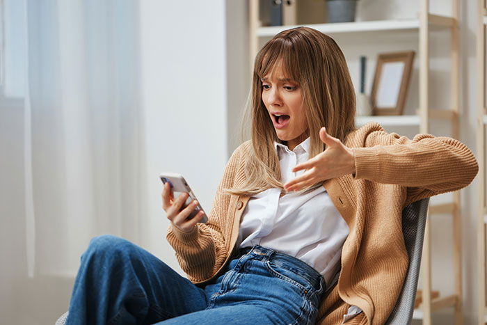 A woman sitting in a chair, looking shocked and upset as she holds her phone, reacting to something on the screen, likely related to the Instagram exposure of the stolen perfumes. A woman sitting in a chair, looking shocked and upset as she holds her phone, reacting to something on the screen, likely related to the Instagram exposure of the stolen perfumes.