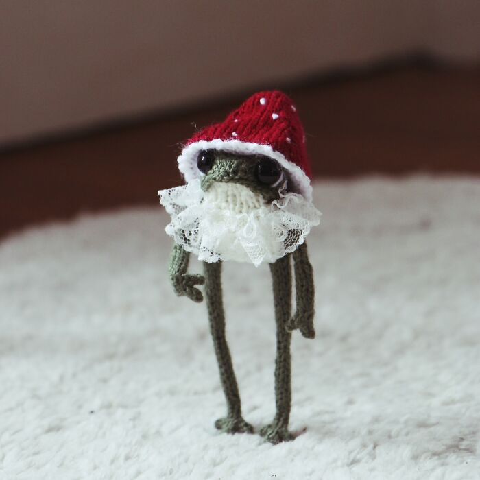 Miniature knitted frog in Santa hat with lace beard, on a soft white surface.